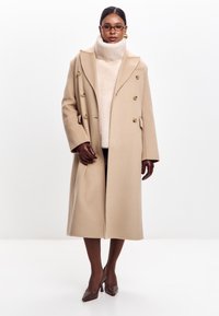 Beige vlněný dvouřadý kabát se zlatými knoflíky, vysokým límcem a oversized střihem; nošený přes krémový rolák, doplněný hnědými rukavicemi a lodičkami.