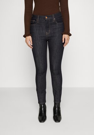 Vrouw in donkere skinny jeans, zwarte enkellaarzen, en een bruine trui met lange mouwen, staand tegen een effen lichte achtergrond.