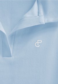 Ljusblå kragblus av ribbad tyg, med en vit broderad logotyp på bröstet och en mjuk, slät textur.