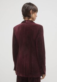 Blazer in velluto bordeaux con superficie testurizzata e lucentezza sottile, dotato di colletto a revers e vestibilità sartoriale.