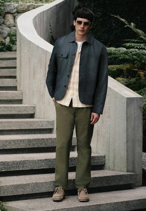 Junger Mann mit Sonnenbrille, grauer Jacke, beigem Hemd, grüner Hose und beigen Schuhen steht auf einer Außentreppe aus Beton in der Nähe von Pflanzen.