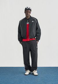 Νεαρός άνδρας που φοράει μαύρο μπουφάν Adidas, μαύρο παντελόνι φόρμας Adidas, κόκκινο πουκάμισο, λευκά αθλητικά παπούτσια και ασπρόμαυρο καπέλο, στέκεται σε μπλε χαλί.