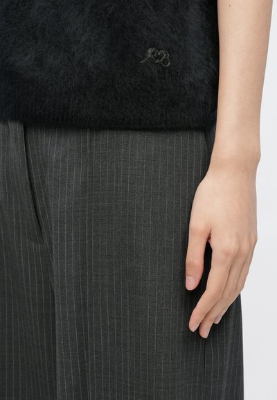 Sort, blød strikket sweater med diskret logodetalje, kombineret med grå bukser med nålestriber. Hånden hviler let på bukserne.