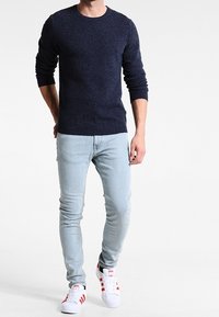 Homme portant un pull texturé bleu marine, un jean skinny bleu clair et des baskets blanches avec des bandes rouges, marchant sur un fond blanc uni.