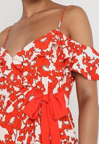 Röd blommig klänning med avklippta volanger vid axlarna, en figursydd midja och en knytning i midjan. Mjuk tyg med en vit bakgrund.