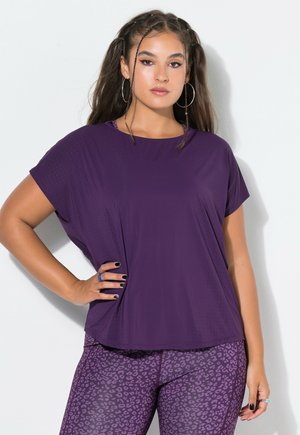 Donna con capelli lunghi indossa una maglia viola larga e leggings viola con stampa leopardata, in piedi con una mano sul fianco su sfondo bianco.