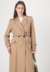 Beige színű gyapjú trench kabát, övvel a deréknál, vállapokkal, két elülső zsebbel és fekete gomb részletekkel. A modellen egy zöld táska van.