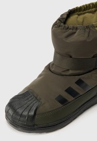 A bota apresenta um upper acolchoado em verde-oliva com uma biqueira e uma tira em borracha preta, decorada com três riscas diagonal pretas. Sola texturizada para melhor aderência.