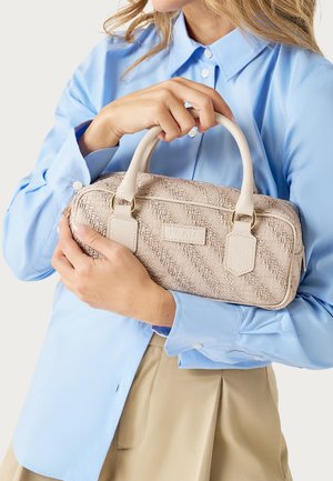 Mujer con una camisa azul claro y una falda beige sosteniendo un bolso beige texturizado con asas redondeadas y herrajes dorados.