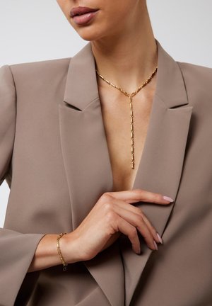 Vrouw in taupe blazer, gouden ketting met verticale hanger en bijpassende gouden armband, hand die voorzichtig de revers van de blazer aanraakt.