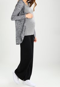 Těhotenský outfit s šedým topem, černými volnými kalhotami a bílými teniskami, doplněný šedým pleteným kardiganem s otevřeným předním dílem.