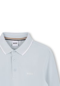 Svijetloplavi polo majica izrađena od teksturirane tkanine. Ima klasični ovratnik, placket s dva gumba i bijeli logo na prsima.