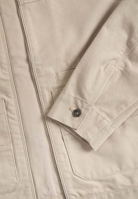 Beige canvas jack met een rits aan de voorkant, knoopmanchetten en opgestikte zakken. De stof heeft een gestructureerde afwerking en strakke lijnen.
