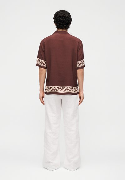 Άντρας με σγουρά σκούρα μαλλιά που φοράει ένα καφέ κοντομάνικο πουκάμισο με κρεμ μπάντες με σχέδια και άσπρο φαρδύ παντελόνι, όρθιος με την πλάτη γυρισμένη.