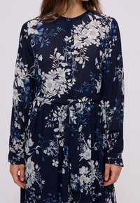 Robe bleu marine à motifs floraux avec un devant boutonné, des manches longues, une ceinture à nouer et un design fluide arborant des motifs floraux bleus et blancs.