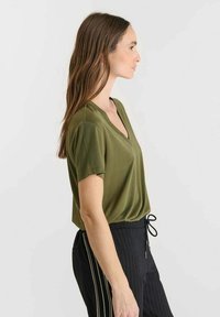 Olijfgroene blouse met korte mouwen en een V-hals, gemaakt van zacht materiaal. Draagt samen met zwarte broek met een streepjespatroon en een trekkoord in de taille.