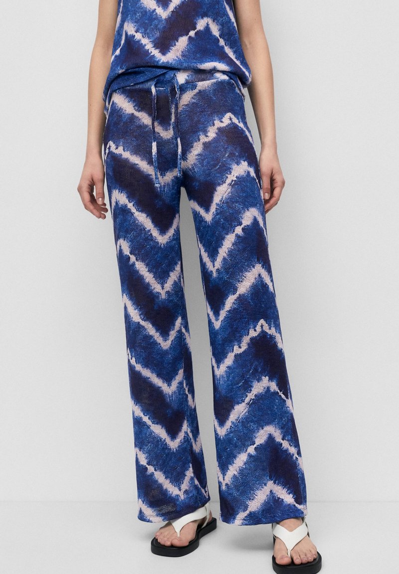 Pantalon à jambes larges avec motif tie-dye bleu et blanc, taille à cordon de serrage, porté avec des sandales blanches à lanières sur une personne debout.