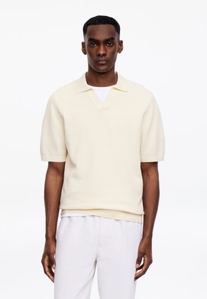 Homme portant un polo à manches courtes crème superposé à un t-shirt blanc et un pantalon blanc, debout devant un fond blanc uni.