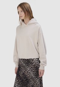 Vrouw die een beige hoodie draagt met de tekst "True" op de schouder en een rok met luipaardprint, staand tegen een effen lichte achtergrond.