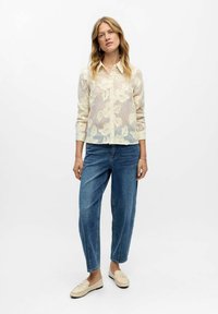 Gennemsigtig beige skjorte med blomsterbroderi, knaplukning foran, lange ærmer; parret med lyseblå jeans og lyse loafers.