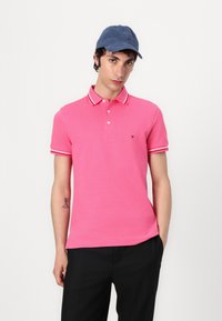 Rosa polo-shirt med kontrasterande vita ränder på kragen och ärmarna, i kombination med en marinblå keps. Kortärmad och med en figurnära design.