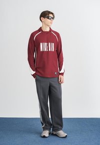 Camisa de manga longa numa cor bordô com detalhes brancos e impressão "NIKE AIR". Calças largas cinzas com riscas laterais. Ténis cinzas.