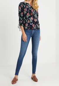 Blommönstrad, marinblå långärmad topp med knytningar på ärmarna, parat med figursydda blåjeans och bruna slip-on skor.