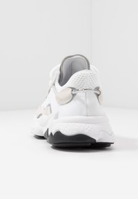 Zapatilla blanca con parte superior texturizada que presenta acentos grises y una suela estampada. Los detalles de cinta de velcro en el talón añaden un diseño funcional.
