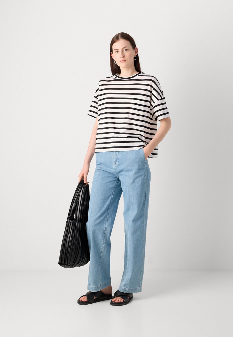 Gestreiftes schwarz-weißes Kurzarmhemd, hellblaue weite Jeans, schwarze flache Sandalen, hält eine strukturierte schwarze Tasche.