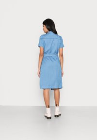 Ljust blå denimskjortklänning med korta ärmar, åtsittande midja och knälång fåll. Bärs med vita ankelskor som har guldaccenter.