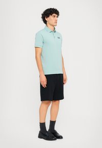 Svetlomodrá polo tričko s krátkymi rukávmi, čierne šortky a čierne mokasíny. Model má kučeravé vlasy a stojí na jednofarebnom pozadí.