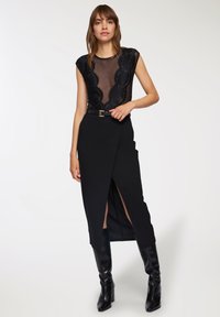 Body nero in pizzo con scollatura a V profonda e maniche a cappuccio, abbinato a una gonna avvolgente nera a vita alta con uno spacco sulla coscia e una cintura coordinata.