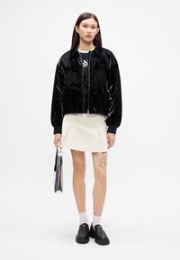 Jachetă bomber neagră și lucioasă, cu fermoar în față și manșete cu nervuri, purtată peste o fustă pliată albă. Pantofi negri slip-on și o geantă argintie completează look-ul.