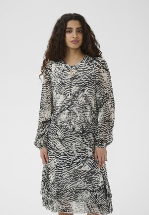 Sieviete ar gariem cirtainiem matiem, tērpusies melnā un baltā abstrakta raksta kleitā ar garām piedurknēm, stāvot pret vienkrāsainu gaišu fonu.