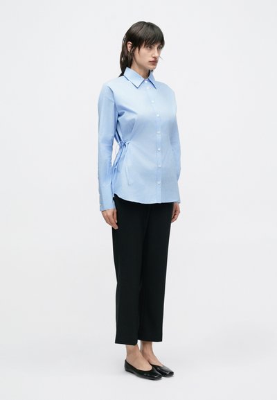 Ανοιχτό μπλε πουκάμισο με κουμπιά, γιακά, πλαϊνές κορδέλες και μακριά μανίκια, σε συνδυασμό με μαύρο παντελόνι ίσιας γραμμής και μαύρες μπαλαρίνες.