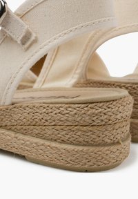 Beige canvas sandalen met gevlochten jute platform, afgeronde rand en een gestructureerde afwerking; voorzien van een veilige strap met metalen hardware.