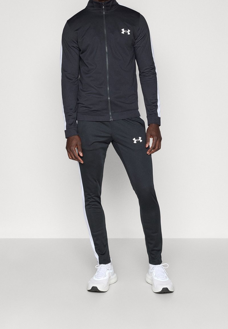 Homme portant un survêtement noir Under Armour avec des bandes blanches sur les manches et les jambes, associé à des baskets blanches, debout devant un fond blanc.
