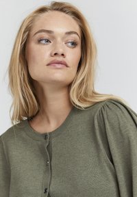 Cardigan lavorato a maglia verde oliva con scollo rotondo e chiusura con bottoni. Presenta maniche a sbuffo e una texture liscia e morbida.