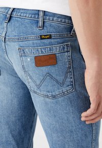 Jeans de ganga azul claro com bolso traseiro visível, apresentando um patch de couro com o logótipo Wrangler e uma pequena etiqueta de tecido Wrangler.