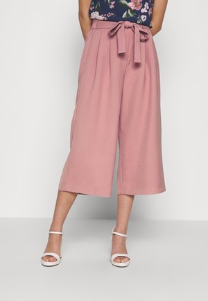 Helle rosa Culottes mit Falten, einem Bindegürtel an der Taille und einem fließenden, strukturierten Stoff, der bis zur Mitte der Waden reicht. Kombiniert mit weißen Sandalen.