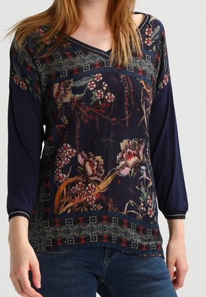 Femme portant un haut à manches longues bleu marine avec des motifs floraux et géométriques, associé à un jean bleu, visible des épaules aux hanches.
