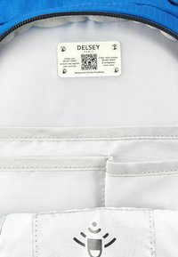 Interior de maleta Delsey con forro de tela azul, etiqueta blanca con un código QR y bolsillos gris claro con un logo bordado.