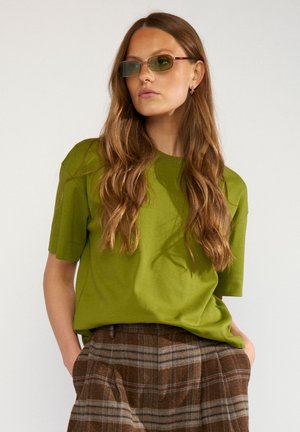 Vrouw met lang golvend haar, gekleed in een olijfgroen T-shirt, bruine geruite broek en smalle groen getinte zonnebril, poseert met handen in de zakken.