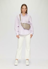 Beige schoudertas met een gladde textuur, ritssluiting en verstelbare schouderband. Draagt over een lichtpaarse gestreepte shirt en witte flapbroeken.