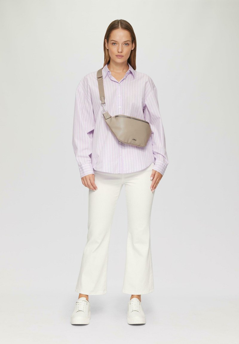 Beige schoudertas met een gladde textuur, ritssluiting en verstelbare schouderband. Draagt over een lichtpaarse gestreepte shirt en witte flapbroeken.
