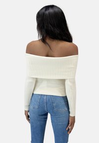 Creamfarbener, schulterfreier, gerippter Strickpullover mit langen Ärmeln und tailliertem Schnitt, kombiniert mit blauen Jeans.