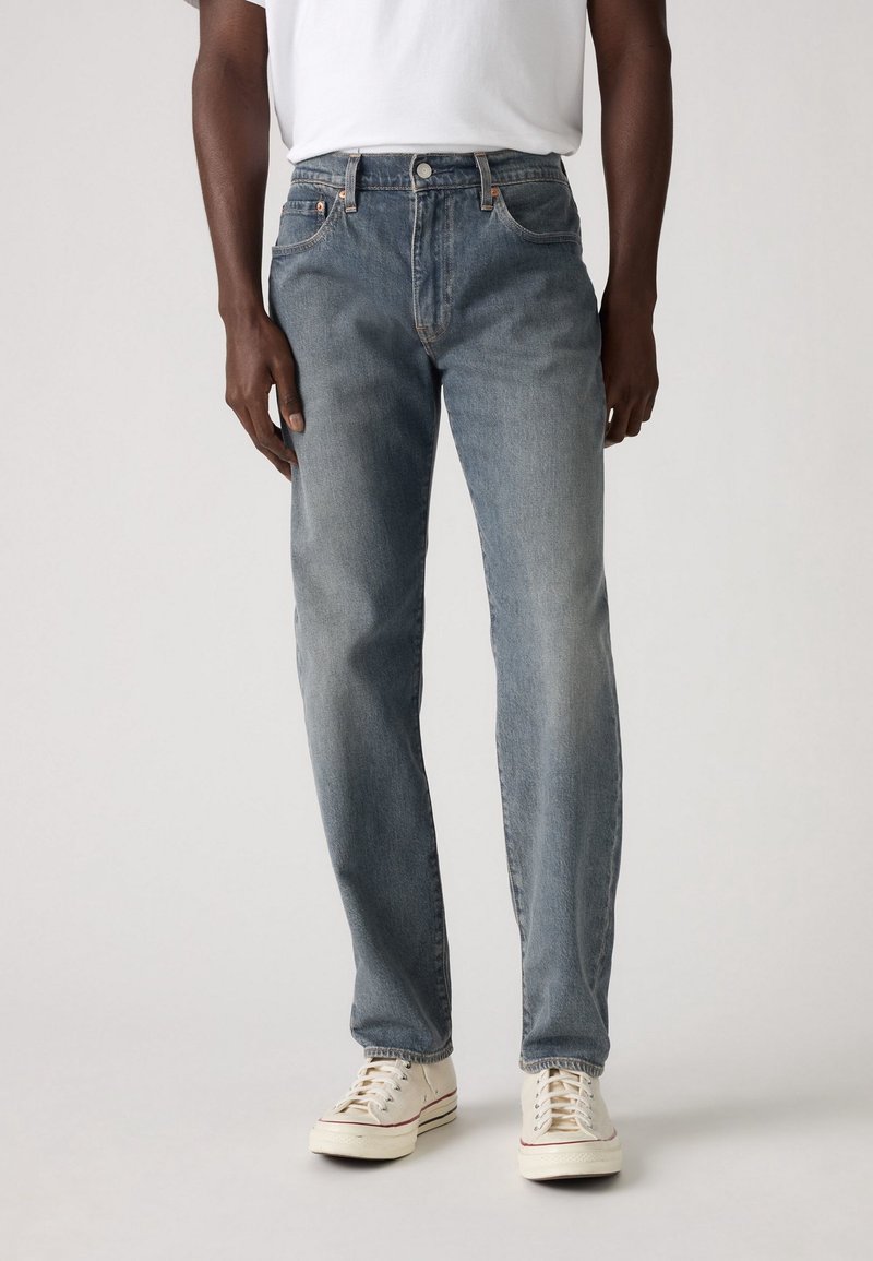 Homme portant un jean droit bleu délavé, des baskets blanches et un t-shirt blanc, debout devant un fond clair uni.