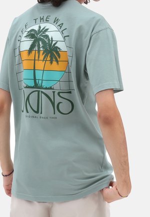 Personne portant un t-shirt bleu clair avec un motif coloré de palmier et le texte "Off The Wall Vans Original Since 1966" dans le dos.