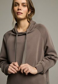 Brązowa, lekka bluza z kapturem o teksturowanym wykończeniu, z długimi rękawami i sznurkami. Posiada luźny krój oraz boczne akcenty.
