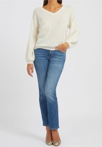 Suéter de punto color crema con escote en V y mangas acampanadas, combinado con jeans azules y tacones altos con estampado de serpiente.